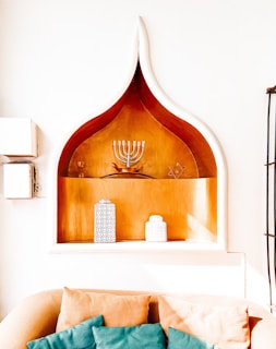 A shelf with a distinct pointed arch design is built into a wall. The shelf features a menorah and a Star of David, indicating Jewish cultural elements. Two decorative canisters with intricate patterns are placed on the lower part of the shelf. In front of the shelf is a beige sofa featuring several teal and beige cushions.