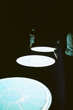 Several illuminated circular tables or platforms stand out against a dark background, with a person partially visible near them, wearing a short dress and possibly a mask.