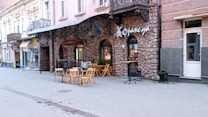 A quiet, cobblestone street lined with pastel-colored buildings. The facade of one building features a rustic stone design and outdoor dining area with wooden tables and chairs. There is a sign with Cyrillic letters above the entrance. A flag is hanging above the window, and several storefronts with awnings and signs are visible further down the street.