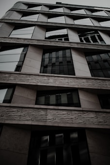 A modern building facade with a geometric design, featuring a series of large windows and textured patterns on the exterior walls. The architectural style looks contemporary, with angled lines and a mix of materials such as glass and concrete.