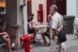 A group of people are gathered outdoors, engaging in a board game on a small table. The surrounding area includes a red fire hydrant, green garbage bins, and chairs. The individuals are casually dressed, and some are wearing masks, indicating health precautions. The setting is urban, with details like a wall-mounted fire hose box and a stack of items on a nearby surface.