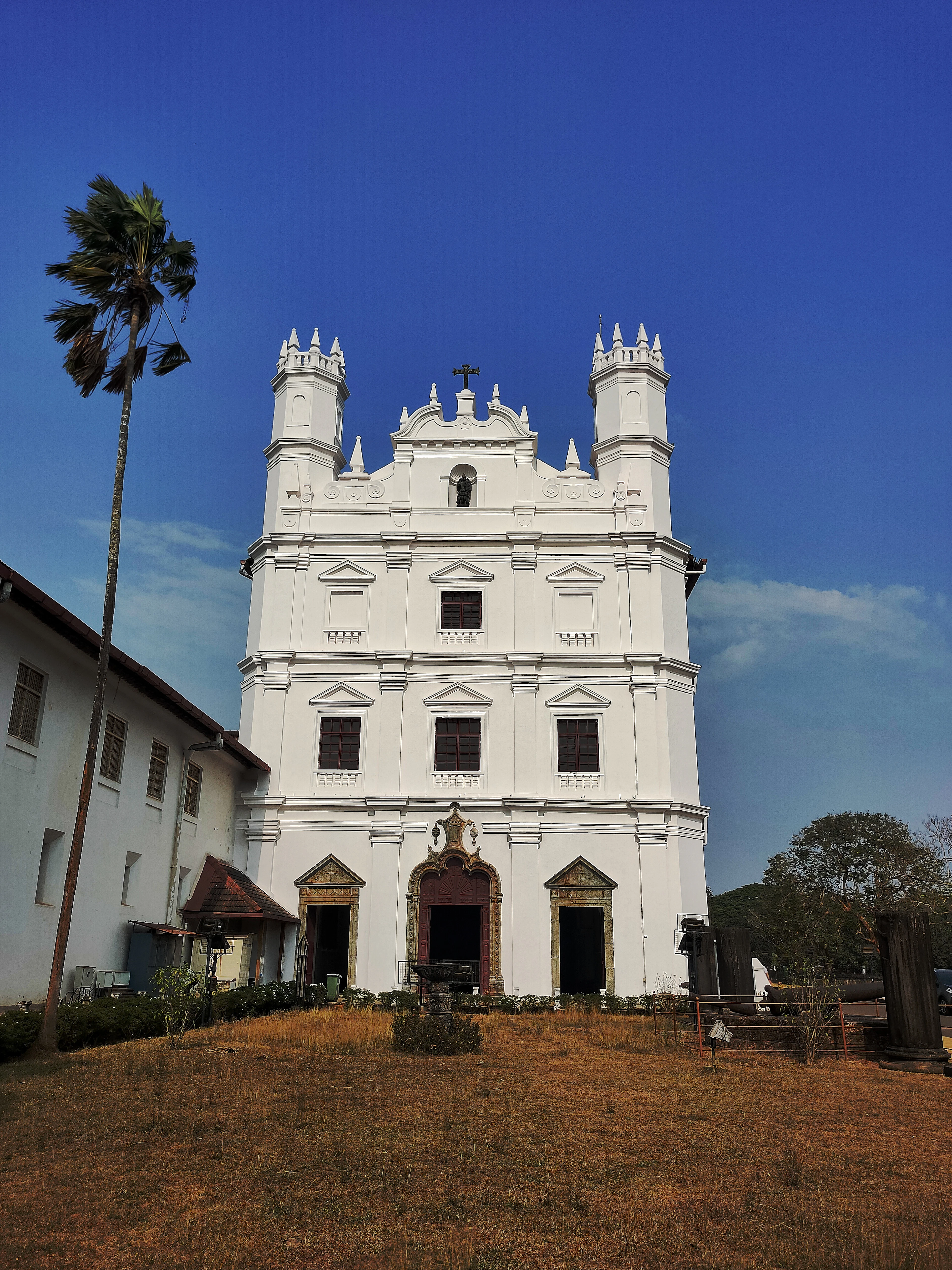 A whitewashed colonial-era church stands beside a palm tree under a bright blue sky in Goa.
