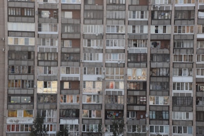A multi-story residential building with a grid of small windows and balconies, each showing different levels of upkeep. The exterior is composed of various materials and colors, including browns, whites, and grays. Some balconies are enclosed, and others are open, with signs of daily life like laundry and decorations visible.