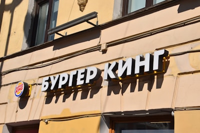 A retail facade with a sign in Cyrillic script indicating a fast-food chain. The letters are bold and white, prominently displayed against a yellow background. A smaller round logo featuring red and blue colors is visible on the left side.