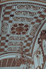 An ornate ceiling with intricate gold patterns and detailed plasterwork, featuring a large, elaborate chandelier hanging from the center. The ceiling design includes symmetrical floral and geometric motifs with a luxurious and classical style.