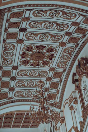 An ornate ceiling with intricate gold patterns and detailed plasterwork, featuring a large, elaborate chandelier hanging from the center. The ceiling design includes symmetrical floral and geometric motifs with a luxurious and classical style.