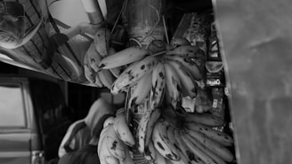 A bunch of ripe bananas hangs prominently, with some dark spots indicating ripeness. They are placed near a vehicle, and various packaged snacks and products are partly visible in the background, suggesting a market or roadside stall setting. A person is seen with their back turned, partially obscured.