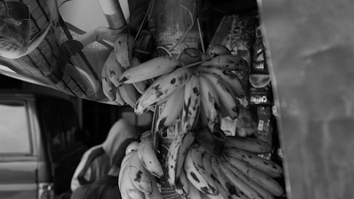 A bunch of ripe bananas hangs prominently, with some dark spots indicating ripeness. They are placed near a vehicle, and various packaged snacks and products are partly visible in the background, suggesting a market or roadside stall setting. A person is seen with their back turned, partially obscured.