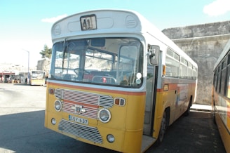 A vintage bus with a yellow and orange exterior is parked in an outdoor setting. The bus has a prominent number 41 displayed at the front and several windows, with some visible reflection on the glass. It is situated next to a stone wall and another bus is partially visible on the right. The background shows a small bus station with people and a few trees.