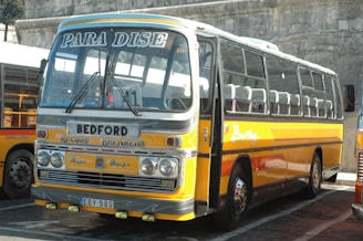 A vintage yellow and orange bus with 'PARA DISE' written on the front above the windshield. The bus has several distinctive markings including 'BEDFORD', 'RECORD BREAKERS', and other decals. It is parked in a lot next to another similar bus. The bus has a classic design with large windows and a boxy structure.