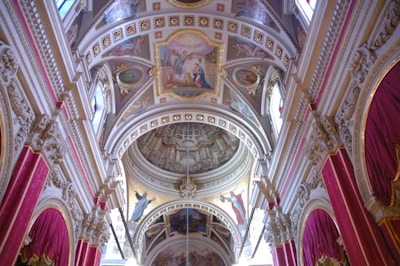 The interior of a grand architectural space is adorned with intricate frescoes and detailed ornamentation. Arched ceilings feature rich decorations with religious paintings, surrounded by ornate moldings and gold accents. Vibrant red curtains hang from the walls, complementing the luxurious design.