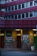 A building facade with multiple windows arranged in a grid pattern, featuring an entrance to a store called 'Reál Pont' in the lower section. The doors are red with signs indicating entry and exit. The walls have graffiti and posters with food images on them. The lighting is dim, suggesting evening or night time.