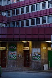 A building facade with multiple windows arranged in a grid pattern, featuring an entrance to a store called 'Reál Pont' in the lower section. The doors are red with signs indicating entry and exit. The walls have graffiti and posters with food images on them. The lighting is dim, suggesting evening or night time.