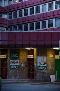 A building facade with multiple windows arranged in a grid pattern, featuring an entrance to a store called 'Reál Pont' in the lower section. The doors are red with signs indicating entry and exit. The walls have graffiti and posters with food images on them. The lighting is dim, suggesting evening or night time.