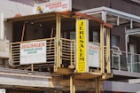 A vintage-style building facade with prominent signs displaying the name 'Jerusalem Sheshkabab House'. The signage includes bold, colorful lettering against a yellow background and faded white wall. There are railings and windows visible, along with an awning providing shade. The overall urban setting suggests a middle-eastern restaurant.