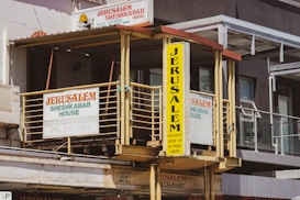 A vintage-style building facade with prominent signs displaying the name 'Jerusalem Sheshkabab House'. The signage includes bold, colorful lettering against a yellow background and faded white wall. There are railings and windows visible, along with an awning providing shade. The overall urban setting suggests a middle-eastern restaurant.