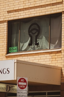 A building facade with a large window displaying a black symbol resembling an eye. The words 'ESCAPE ROOMS' are illuminated in green neon on the window. Below, a 'NO ENTRY' sign is posted with 'BICYCLES EXCEPTED' underneath. The building is constructed with tan bricks.