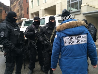 A group of individuals dressed in tactical gear and black uniforms stand in a formation, appearing to be security or law enforcement personnel. They are engaged in conversation with someone wearing a blue winter jacket with a fur-lined hood, displaying the text 'Igor Zhilkin Production' on the back. The setting appears to be an urban area with buildings in the background.