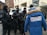A group of individuals dressed in tactical gear and black uniforms stand in a formation, appearing to be security or law enforcement personnel. They are engaged in conversation with someone wearing a blue winter jacket with a fur-lined hood, displaying the text 'Igor Zhilkin Production' on the back. The setting appears to be an urban area with buildings in the background.
