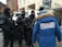 A group of individuals dressed in tactical gear and black uniforms stand in a formation, appearing to be security or law enforcement personnel. They are engaged in conversation with someone wearing a blue winter jacket with a fur-lined hood, displaying the text 'Igor Zhilkin Production' on the back. The setting appears to be an urban area with buildings in the background.
