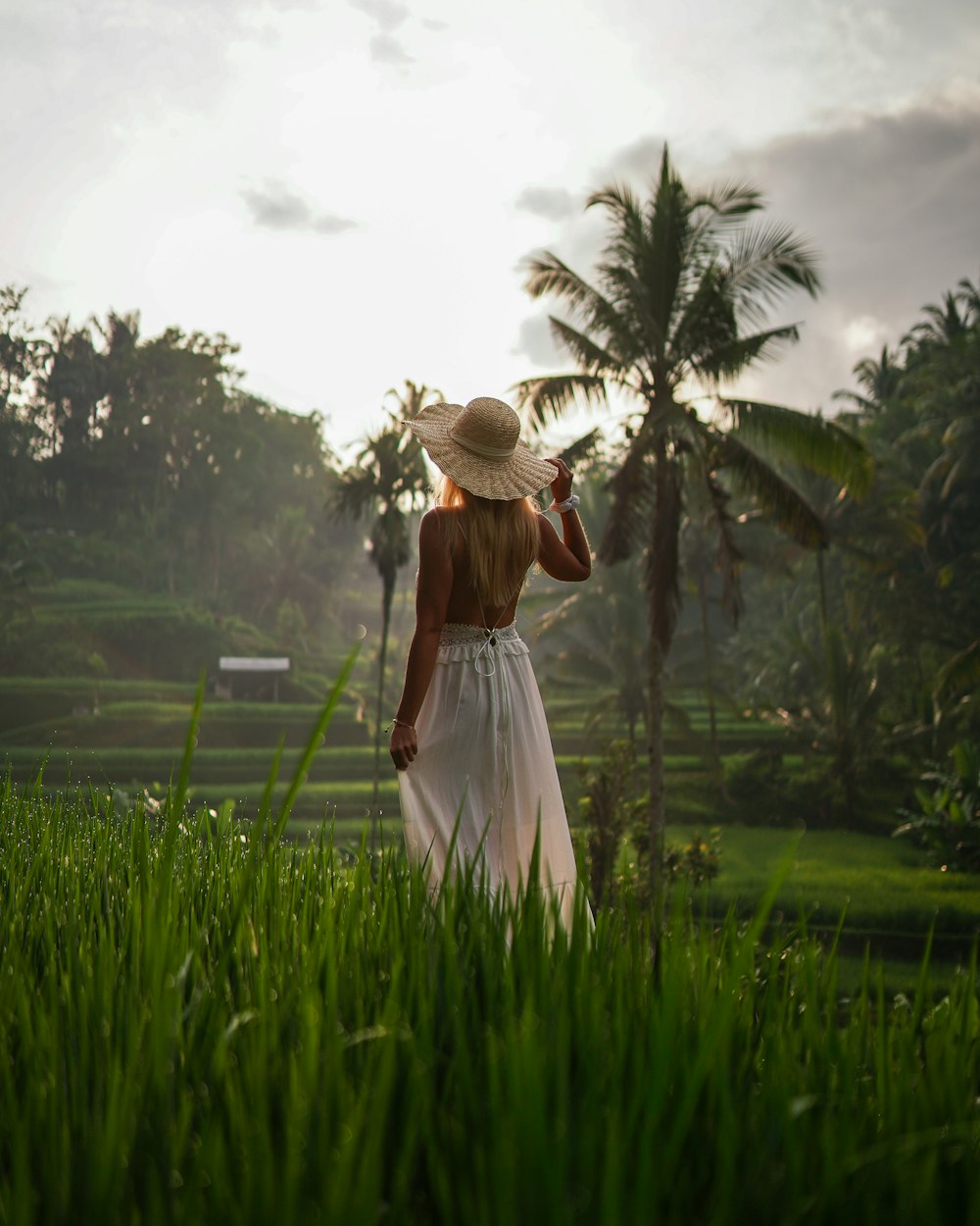 a woman in a white dress and hat walking through a lush green field