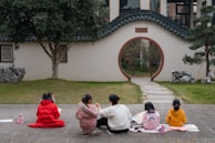 A group of children and an adult are sitting on the pavement in front of a traditional Asian-style garden with a circular moon gate. They are engaged in an activity, possibly drawing or studying, as there are papers and water bottles around them. The garden features neatly trimmed grass, rocks, and a few trees.