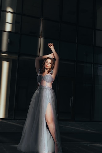 Softly lit scene of a woman in an elegant evening gown, poised by a large window at dusk.