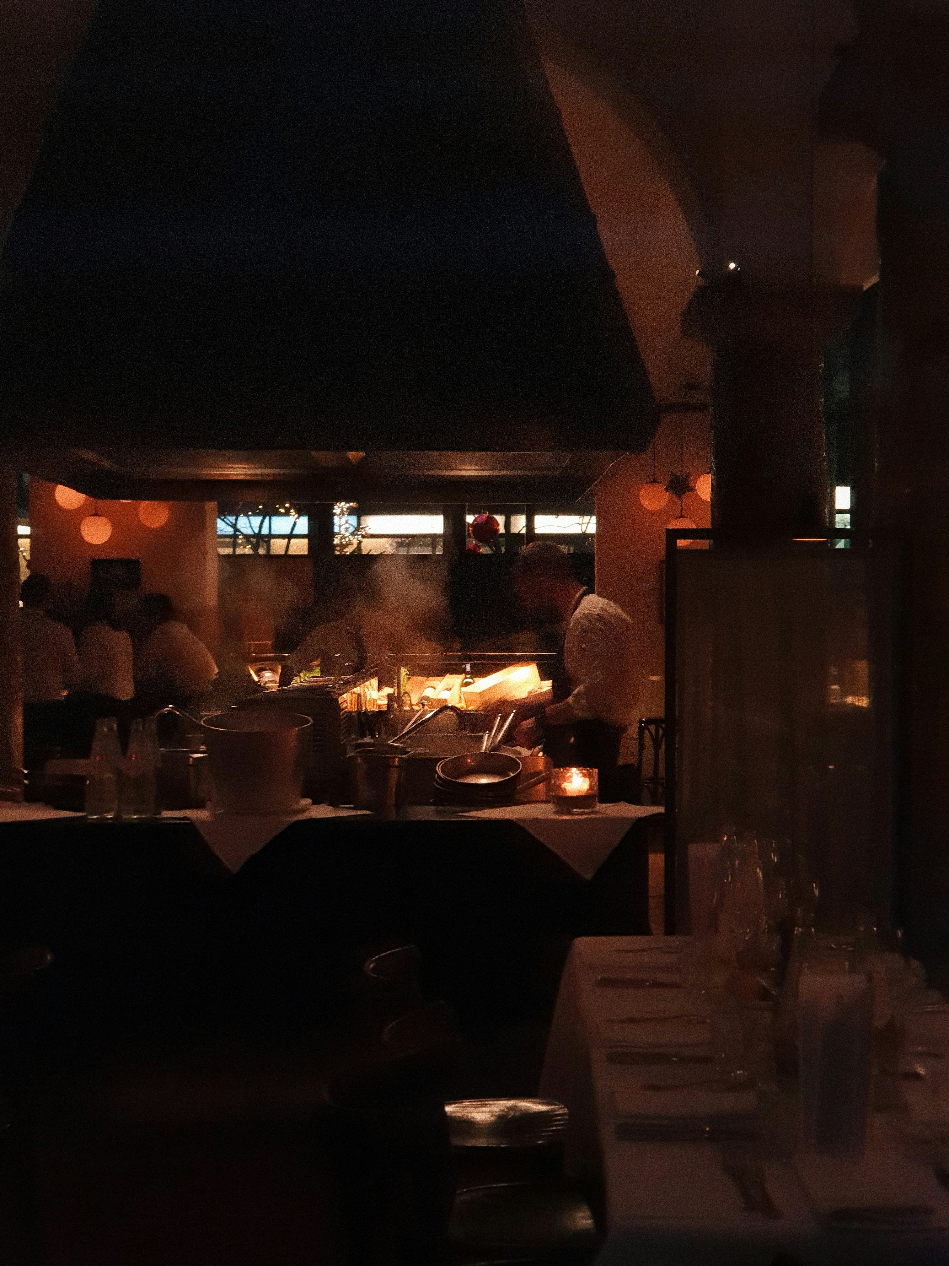 Restaurant kitchen at night, chefs working under dim amber lighting, dark steel counters, moody shadows