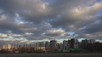 High-rise apartment buildings under a partly cloudy sky with dense, dark clouds casting shadows on the structures. The urban skyline is contrasted with small green-roofed houses in the foreground and a barren landscape.