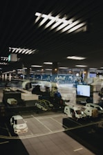 A modern airport scene shows a parked airplane with ground service vehicles nearby. The setting appears to be nighttime with the interior airport lights reflecting on a large glass window, adding depth and complexity to the image.