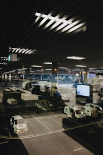 A modern airport scene shows a parked airplane with ground service vehicles nearby. The setting appears to be nighttime with the interior airport lights reflecting on a large glass window, adding depth and complexity to the image.