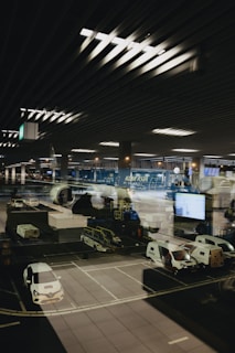 A modern airport scene shows a parked airplane with ground service vehicles nearby. The setting appears to be nighttime with the interior airport lights reflecting on a large glass window, adding depth and complexity to the image.
