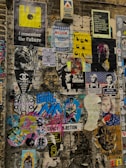 A vibrant collage of various posters and stickers on a brick wall, featuring eclectic imagery and text. The wall displays a mix of retro graphics, such as a yellow floppy disk, along with contemporary art and photographs. There is a mix of satirical, pop culture, and surreal references, including images of people, cartoon characters, and abstract designs. The poster colors and themes vary widely, creating an urban, chaotic feel.