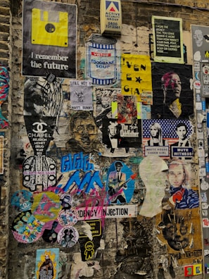 A vibrant collage of various posters and stickers on a brick wall, featuring eclectic imagery and text. The wall displays a mix of retro graphics, such as a yellow floppy disk, along with contemporary art and photographs. There is a mix of satirical, pop culture, and surreal references, including images of people, cartoon characters, and abstract designs. The poster colors and themes vary widely, creating an urban, chaotic feel.