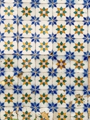 An ornate pattern of ceramic tiles featuring star-like geometric designs. Each tile displays a central star shape with alternating gold and green diamond shapes and blue corner pieces. Some tiles show signs of wear and aging, including cracks and discoloration.