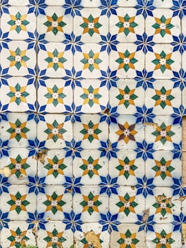 An ornate pattern of ceramic tiles featuring star-like geometric designs. Each tile displays a central star shape with alternating gold and green diamond shapes and blue corner pieces. Some tiles show signs of wear and aging, including cracks and discoloration.