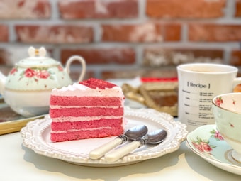 A slice of pink layered cake sits on a ornate white plate, accompanied by two spoons with white handles. In the background, a floral teapot and a cup with the text 'Recipe' are visible, all set on a table with a brick wall backdrop.