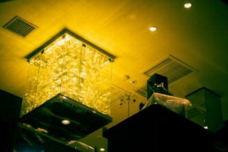 A ceiling features intricate lighting with a large, illuminated glass installation emitting a yellow-green glow. The room's modern interior includes ceiling vents, recessed lighting, and a mixture of geometric shapes.