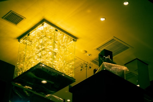 A ceiling features intricate lighting with a large, illuminated glass installation emitting a yellow-green glow. The room's modern interior includes ceiling vents, recessed lighting, and a mixture of geometric shapes.