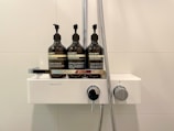 Three large, dark bottles with pump dispensers are arranged on a modern, white shower fixture. Each bottle is labeled with text, likely indicating different types of bath products such as shampoo, conditioner, and shower gel. A metal hose is attached to the fixture, suggesting a shower setting.