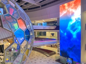 A modern interior featuring a large digital display with a vibrant abstract pattern of red, blue, and white colors. Adjacent to it, there is a structure composed of hexagonal panels, each reflecting a variety of hues. The space is open with multiple levels visible, including staircases and glass railings. The floor pattern enhances the contemporary aesthetic.