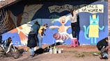 A group of people are painting a colorful mural on a wall. The mural features a large bird, a human-like figure with raised arms, and a cat-like creature wearing a yellow dress and crown. There are paint cans and other painting supplies scattered on the ground. A bicycle is parked nearby, and the slogan 'Women for Peace, Peace for Women' is visible in bold letters.