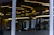 Abstract architectural design featuring reflective glass surfaces with curved yellow lighting accents. Vertical lines from support pillars intersect with curved light patterns, creating a modern and artistic visual. The interior appears sophisticated, with a mix of metallic and glass elements adding depth through reflections.