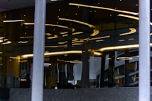 Abstract architectural design featuring reflective glass surfaces with curved yellow lighting accents. Vertical lines from support pillars intersect with curved light patterns, creating a modern and artistic visual. The interior appears sophisticated, with a mix of metallic and glass elements adding depth through reflections.