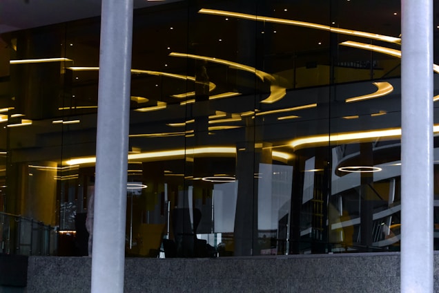 Abstract architectural design featuring reflective glass surfaces with curved yellow lighting accents. Vertical lines from support pillars intersect with curved light patterns, creating a modern and artistic visual. The interior appears sophisticated, with a mix of metallic and glass elements adding depth through reflections.