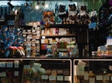 A dimly lit candy and chocolate shop with a variety of sweets displayed on shelves and counters. The shelves are filled with packaged chocolates and candies, some with colorful wrappings and labels. Decorative items like Easter eggs and rabbit figurines are present, along with price tags. Products are arranged neatly, with larger wrapped items like gift packs visible. Overhead lighting casts a warm ambiance over the assortment of confections.