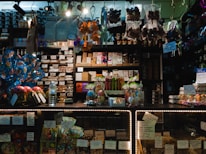 A dimly lit candy and chocolate shop with a variety of sweets displayed on shelves and counters. The shelves are filled with packaged chocolates and candies, some with colorful wrappings and labels. Decorative items like Easter eggs and rabbit figurines are present, along with price tags. Products are arranged neatly, with larger wrapped items like gift packs visible. Overhead lighting casts a warm ambiance over the assortment of confections.