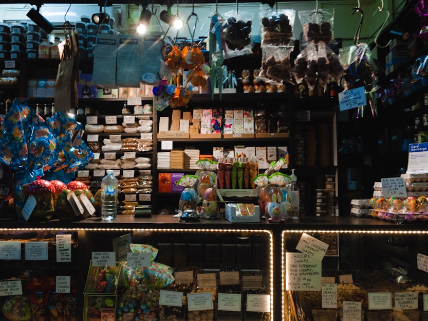 A dimly lit candy and chocolate shop with a variety of sweets displayed on shelves and counters. The shelves are filled with packaged chocolates and candies, some with colorful wrappings and labels. Decorative items like Easter eggs and rabbit figurines are present, along with price tags. Products are arranged neatly, with larger wrapped items like gift packs visible. Overhead lighting casts a warm ambiance over the assortment of confections.