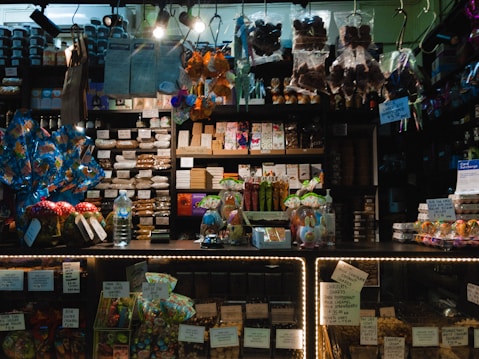 A dimly lit candy and chocolate shop with a variety of sweets displayed on shelves and counters. The shelves are filled with packaged chocolates and candies, some with colorful wrappings and labels. Decorative items like Easter eggs and rabbit figurines are present, along with price tags. Products are arranged neatly, with larger wrapped items like gift packs visible. Overhead lighting casts a warm ambiance over the assortment of confections.