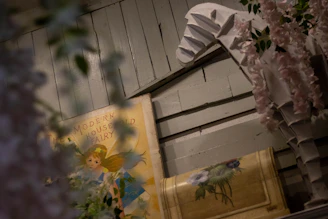 A vintage-style book titled 'The Modern Household Fairy' is partially obscured by floral arrangements, set against a backdrop of wooden paneling. A sculpture resembling a horse is adorned with pink flowers.
